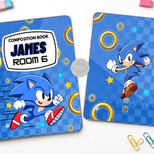 Puede incluir: Dos cuadernos de composición con temática de Sonic el Erizo azul, con anillos dorados y estrellas amarillas sobre un fondo a cuadros. La portada muestra el texto "COMPOSITION BOOK JAMES ROOM 6" en una etiqueta blanca con borde negro.