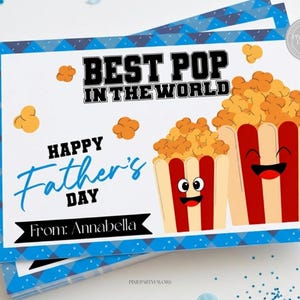 Puede incluir: Una pila de tarjetas del Día del Padre con un borde geométrico azul y blanco. Las tarjetas presentan el texto "BEST POP IN THE WORLD" y "HAPPY Father's DAY". También se muestran dos cubos de palomitas de dibujos animados con caras sonrientes. La tarjeta está firmada "From: Annabella".