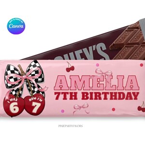 May include: A pink candy bar wrapper with the name "Amelia" and "7th Birthday" in red lettering. The wrapper features a black and white checkered bow, cherries, and the text "I was 6, now I'm 7". A Hershey's bar is in the background.
