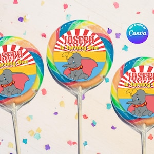 May include: Three lollipops with a cartoon elephant design. Each lollipop has the text "JOSEPH is turning one" and a picture of a gray elephant with a red cape. The lollipops have a rainbow swirl pattern and are on clear sticks.