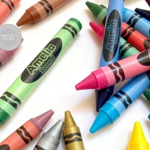 Puede incluir: Una colección de ceras de colores esparcidas sobre una superficie blanca. Cada cera presenta una etiqueta ovalada negra impresa con el texto Amelia Room 8 en una fuente de color verde claro. Las ceras aparecen en varios tonos, incluyendo verde, azul, rosa, amarillo, marrón y plata metálica.