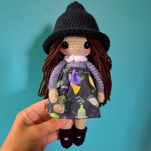 May include: A handmade crochet doll featuring long brown yarn hair, a black pointed hat, and a light purple long-sleeved top with a white collar. The doll wears a black skirt patterned with colorful illustrations of bottles, herbs, and hats.