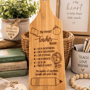 Op de afbeelding: Een lichtgekleurde, peddelvormige snijplank van hout met een in laser gegraveerde tekst The Perfect Teacher Recipe, samen met illustraties van een boek, een appel, een wereldbol en een potlood, plus een lijst met metaforische ingrediënten.