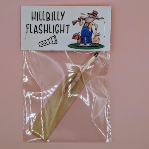 Pode incluir: Um saco de plástico transparente contendo um pau de madeira com um fósforo preso a ele. O saco tem um rótulo branco que diz "Hillbilly Flashlight" com uma imagem de desenho animado de um homem segurando uma arma e um cão.