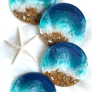 Pode incluir: Quatro bases redondas de resina com tema de praia. Cada base apresenta um design de oceano azul escuro e turquesa com ondas brancas e espumosas e pequenas pedras na parte inferior. Uma estrela do mar branca está ao lado das bases.