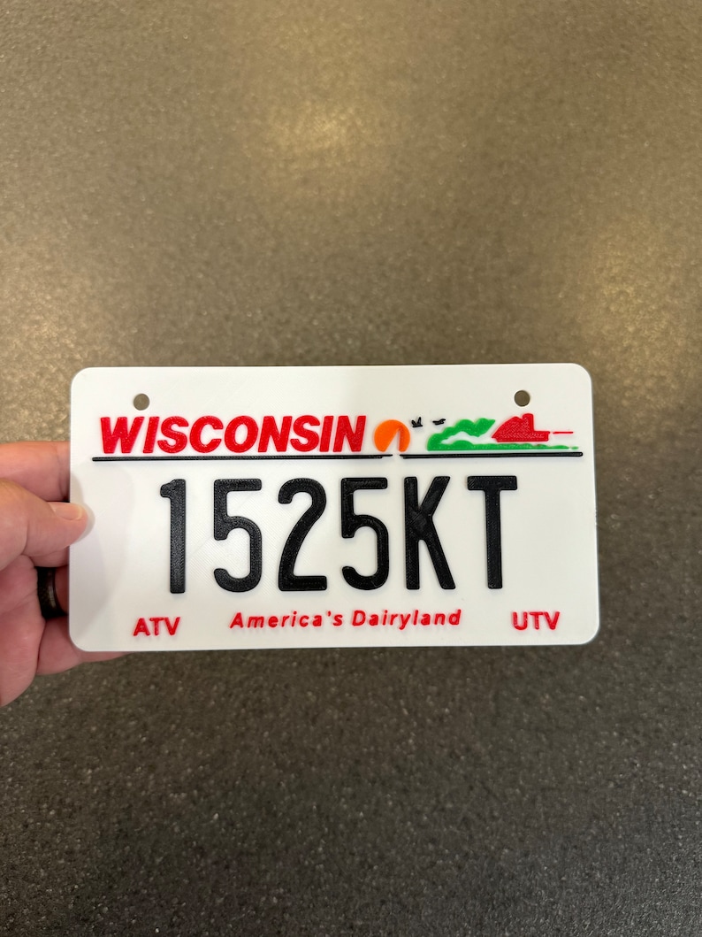 May include: White Wisconsin license plate with red text and a red, orange, and green graphic. The license plate number is 1525KT. The text "America's Dairyland" is printed in red below the number. The words "ATV" and "UTV" are printed in red on either side of the text.