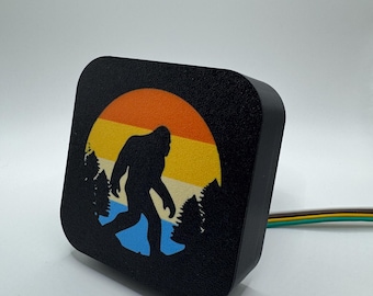 May include: Black square trailer hitch cover featuring a silhouette of Bigfoot walking in front of a sunset. The sunset is orange, yellow, and blue.