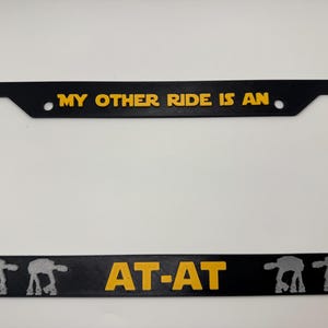 Puede incluir: Marco de matrícula negro con texto amarillo "MY OTHER RIDE IS AN" encima de "AT-AT". El marco tiene un borde negro y presenta siluetas de caminantes AT-AT.