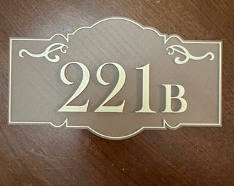 May include: A brown wooden sign with a gold-colored number "221B" on it. The sign has a decorative border.