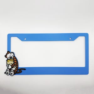 May include: A rectangular blue license plate frame featuring a graphic of a cartoon tiger hugging a small boy on the lower left side. The frame has two mounting holes at the top and a smooth, solid blue finish across the remaining border area.