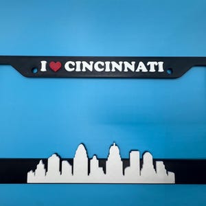 May include: Black license plate frame with the text "I ❤️ CINCINNATI" and a white skyline silhouette. The heart is red. The frame is designed to fit a standard U.S. license plate.