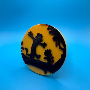 May include: A round, yellow decorative item with a black silhouette of two cartoon characters. One character is jumping with arms outstretched, and the other is running. The background is a bright yellow color.