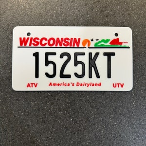 May include: White Wisconsin license plate with red lettering and a sunset graphic. The plate number is 1525KT. The text "America's Dairyland" is printed below the number.