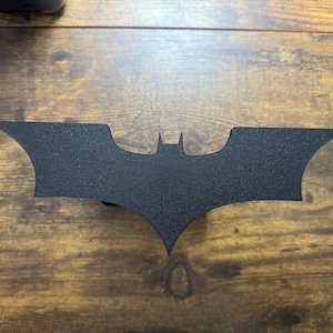 May include: A black metal Batman logo cutout. The logo is in the shape of a bat with a pointed wingspan and a small, pointed head.