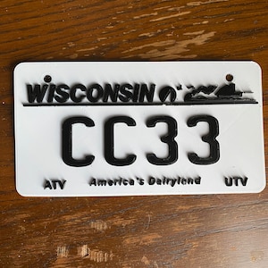 May include: White Wisconsin ATV/UTV license plate with black lettering and a black silhouette of a boat. The plate number is CC33. The text "America's Dairyland" is printed below the number.