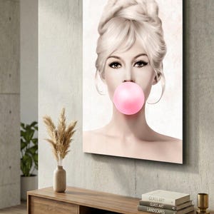 May include: A framed art print featuring a woman with blonde hair styled in an updo, blowing a pink bubble. The artwork is displayed on a gray wall above a wooden cabinet with decorative items. The overall aesthetic is modern and stylish.