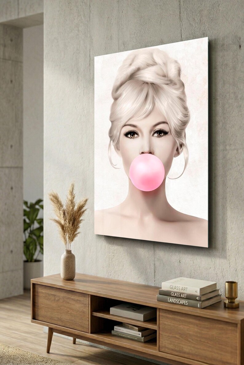 May include: A framed art print featuring a woman with blonde hair styled in an updo, blowing a pink bubble. The artwork is displayed on a gray wall above a wooden cabinet with decorative items. The overall aesthetic is modern and stylish.