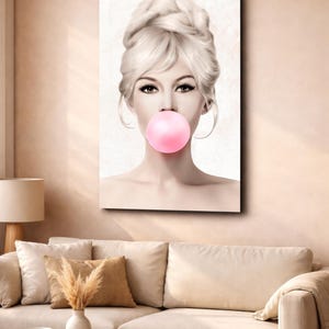 May include: A canvas print featuring a woman with blonde hair styled in an updo, blowing a large pink bubble. The artwork has a vintage aesthetic, with soft lighting and a neutral background. The woman's eyes are emphasized with dark eyeliner.