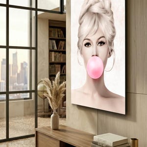 May include: A canvas print featuring a woman with blonde hair and a pink bubble gum bubble. The artwork has a vintage aesthetic, with the woman's face as the focal point. The background is a neutral color, and the image is displayed in a modern interior.