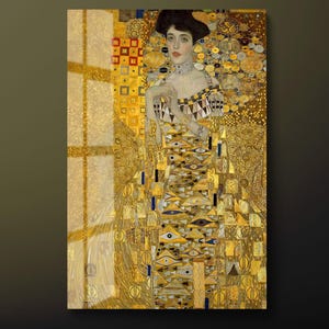 May include: A framed print of a Gustav Klimt painting. The artwork features a woman in a gold dress with geometric patterns and intricate details. The background is filled with gold and yellow decorative elements, creating a rich and ornate aesthetic.