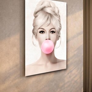 May include: A framed art print featuring a woman with blonde hair in a bouffant hairstyle, blowing a large pink bubble. The artwork has a vintage aesthetic, with soft lighting and a neutral background. The print is displayed on a wall.