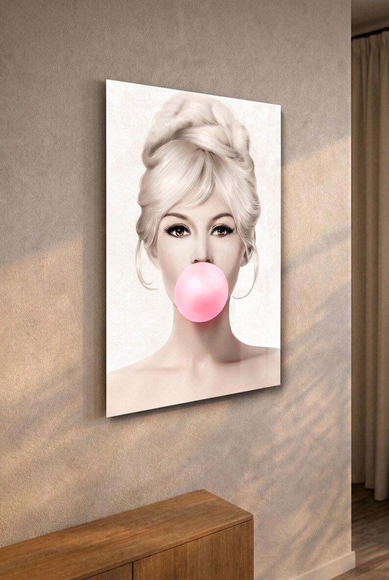 May include: A framed art print featuring a woman with blonde hair in a bouffant hairstyle, blowing a large pink bubble. The artwork has a vintage aesthetic, with soft lighting and a neutral background. The print is displayed on a wall.