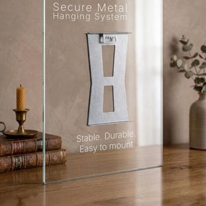 May include: A clear glass panel with a secure metal hanging system. The text on the glass reads "Secure Metal Hanging System" and "Stable. Durable. Easy to mount." The metal bracket is silver. The panel is on a wooden surface.