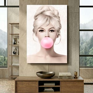 May include: A canvas print featuring a portrait of a woman with blonde hair styled in an updo, blowing a large pink bubblegum bubble. The artwork is displayed on a wall above a wooden cabinet, creating a stylish interior design.