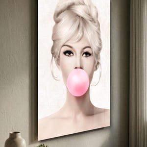 May include: A framed art print featuring a woman with blonde hair styled in an updo, blowing a large pink bubble. The artwork has a soft, pastel color palette and is mounted on a wall. The image is a portrait.