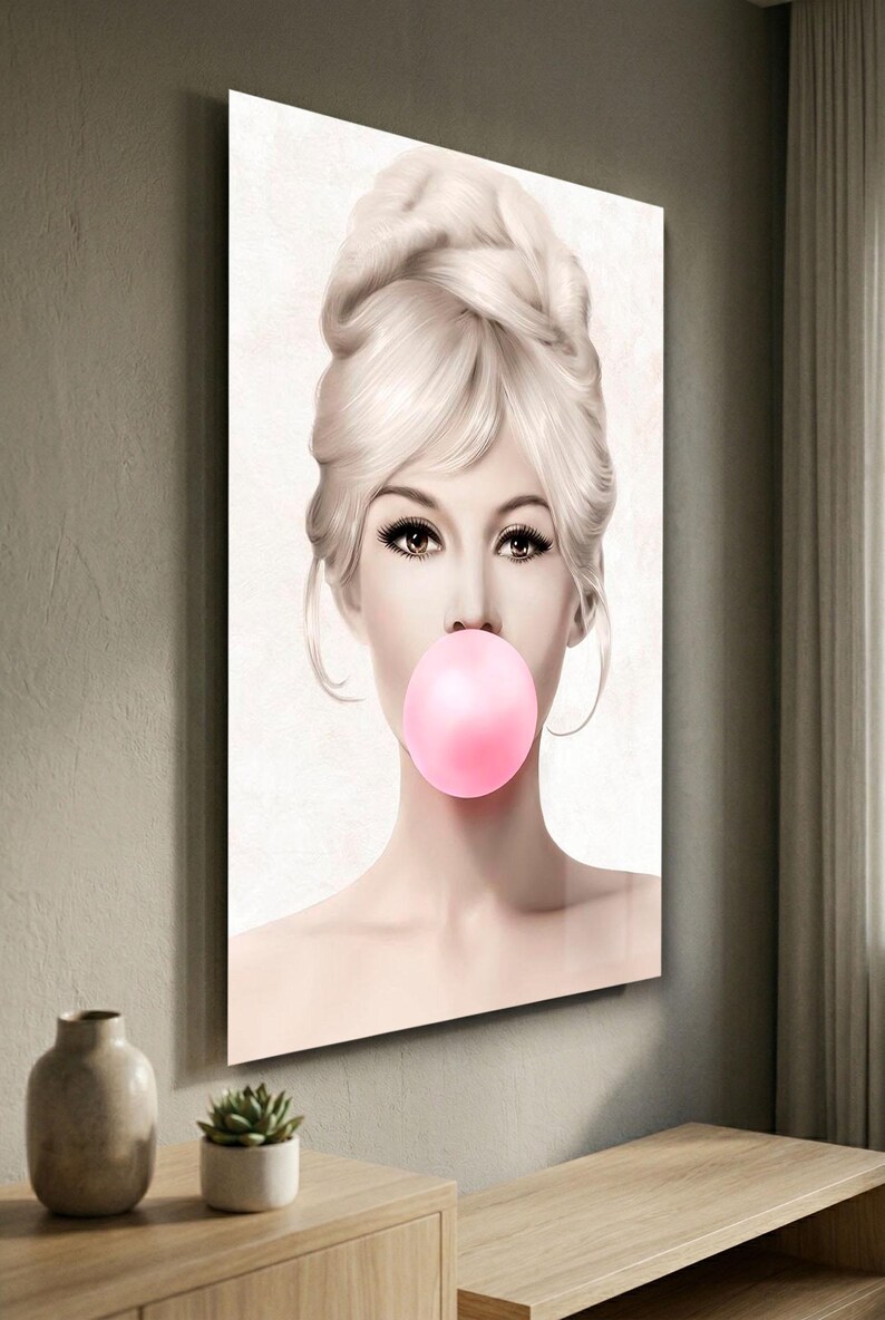 May include: A framed art print featuring a woman with blonde hair styled in an updo, blowing a large pink bubble. The artwork has a soft, pastel color palette and is mounted on a wall. The image is a portrait.