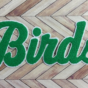 May include: A green chenille patch featuring the word Birds in a cursive script font. The lettering has a textured, fuzzy surface and is outlined with a thick white border, creating a bold and distinct appearance for this decorative applique.