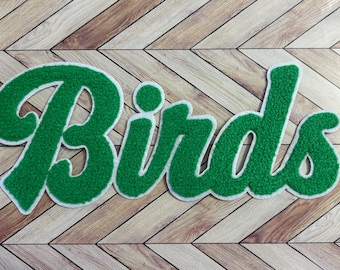 May include: A green chenille patch featuring the word Birds in a cursive script font. The lettering has a textured, fuzzy surface and is outlined with a thick white border, creating a bold and distinct appearance for this decorative applique.