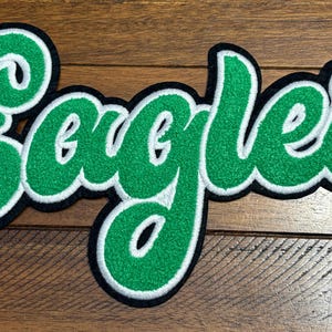 May include: A green and white embroidered patch featuring the word Eagles in a cursive script font. The lettering has a bright green textured center, a thick white border, and a thin black outer edge, creating a bold layered appearance.