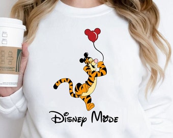 Könnte beinhalten: Weißer Pullover mit einem Cartoon-Bild von Tigger aus Winnie Puuh, der einen roten Ballon mit Mickey-Maus-Ohren hält. Der Text "Disney Mode" ist unter dem Bild gedruckt.
