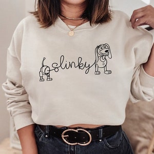 May include: A cream-colored sweatshirt with a black outline of Slinky Dog from the Toy Story franchise. The text "Slinky" is written in cursive above the dog.