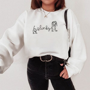 May include: White sweatshirt with a black outline of Slinky Dog from Toy Story. The text "Slinky" is written in cursive above the dog.