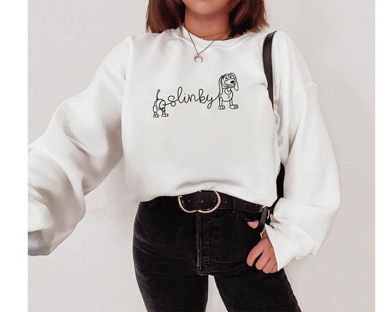 May include: White sweatshirt with a black outline of Slinky Dog from Toy Story. The text "Slinky" is written in cursive above the dog.