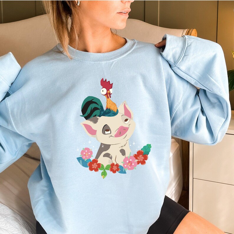 K&ouml;nnte beinhalten: Ein hellblaues Sweatshirt mit einem Cartoon-Bild eines Schweins mit einem Hahn auf dem Kopf. Das Schwein ist von Blumen und Bl&auml;ttern umgeben.