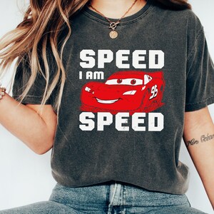 Puede incluir: Una camiseta gris oscuro con un coche de carreras rojo de dibujos animados y el texto "SPEED I AM SPEED" en blanco.