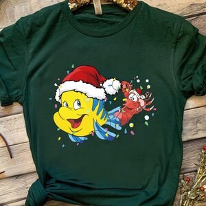 Puede incluir: Una camiseta verde con una imagen de dibujos animados de un pez amarillo con un gorro de Pap&aacute; Noel y una langosta roja con un gorro de Pap&aacute; Noel. El pez y la langosta est&aacute;n rodeados de luces navide&ntilde;as de colores.