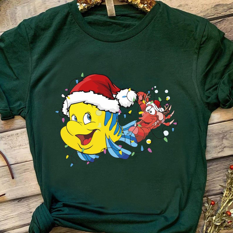 Puede incluir: Una camiseta verde con una imagen de dibujos animados de un pez amarillo con un gorro de Pap&aacute; Noel y una langosta roja con un gorro de Pap&aacute; Noel. El pez y la langosta est&aacute;n rodeados de luces navide&ntilde;as de colores.