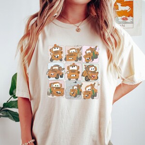 Puede incluir: Una camiseta blanca con una imagen de dibujos animados de una gr&uacute;a de remolque con diferentes expresiones faciales. La gr&uacute;a de remolque es marr&oacute;n y tiene un parachoques verde.