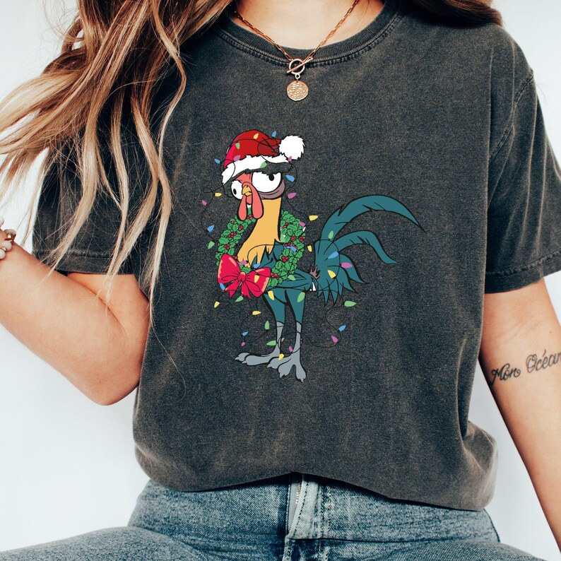 May include: A dark gray t-shirt with a cartoon rooster wearing a Santa hat and a Christmas wreath. The rooster is surrounded by colorful Christmas lights.