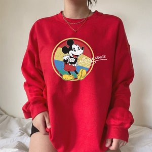 Puede incluir: Sudadera roja con un gr&aacute;fico vintage de Mickey Mouse y el texto "Mickey Mouse" en blanco. El gr&aacute;fico presenta a Mickey Mouse en un c&iacute;rculo amarillo, rojo y azul.