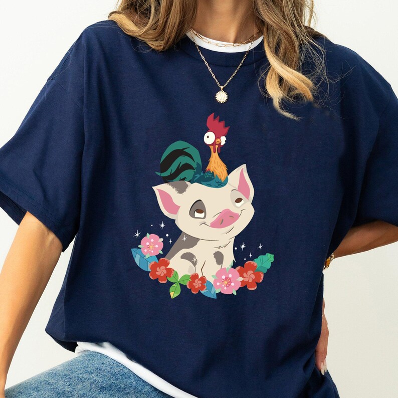 K&ouml;nnte beinhalten: Ein marineblaues T-Shirt mit einem wei&szlig;en Untergrund, das ein Cartoon-Schwein mit einem Hahn auf dem Kopf zeigt. Das Schwein ist wei&szlig; mit schwarzen Flecken und hat eine rosa Nase. Der Hahn ist rot und hat einen gr&uuml;n-blauen Schwanz. Das Design ist von rosa und roten Blumen umgeben.