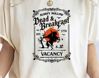 May include: White t-shirt with a black and white graphic design of a sign that says "Sleepy Hollow Dead & Breakfast Est. 1776" with a black silhouette of a headless horseman riding a horse with a pumpkin in his hand. The sign also says "Vacancy Where you're sure to rest in peace."
