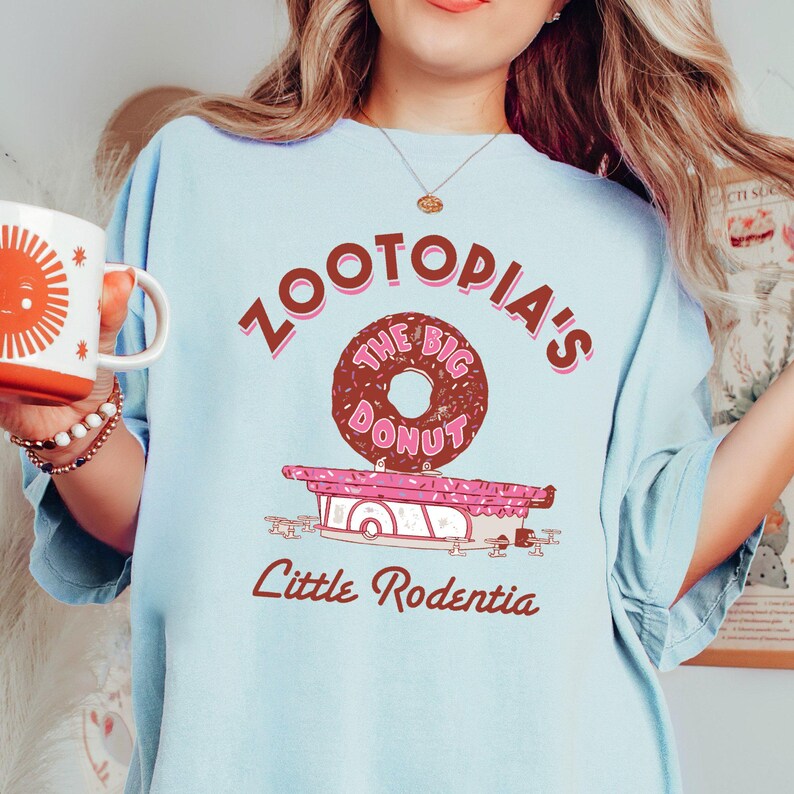 May include: Light blue t-shirt with a pink and brown graphic of a donut shop called "Zootopia's The Big Donut" with the text "Little Rodentia" below.