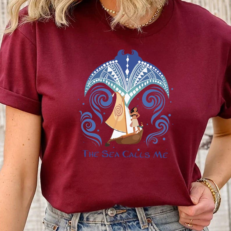 May include: A maroon t-shirt with a graphic of a woman sailing on a boat with a manta ray design above her. The text "The Sea Calls Me" is printed below the graphic.