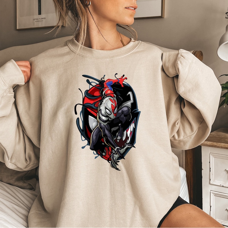 Puede incluir: Una sudadera beige con un estampado gr&aacute;fico de un personaje Venom en blanco y negro con un personaje Spider-Man rojo y azul dentro de la cabeza del personaje Venom.