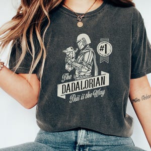 K&ouml;nnte beinhalten: Ein dunkelgraues T-Shirt mit einem Motiv eines Mandalorianer-Charakters, der ein Baby Yoda h&auml;lt. Der Text "The Dadalorian This is the Way" ist auf dem Shirt aufgedruckt. Das Motiv enth&auml;lt auch ein Abzeichen mit der Aufschrift "Certified #1 in the Galaxy".